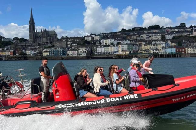 Group on a red speedboat in harbor, town and church in the background under blue sky.